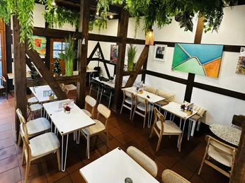 Innenansicht Café Helles Café mit weißen Tischen, Holzelementen, grünen Pflanzen und farbenfrohen Wandbildern.