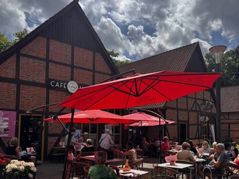 Außenansicht Sommer.jpeg Gäste genießen Kaffee und Kuchen unter roten Sonnenschirmen vor einem Fachwerkhaus im Sommer.