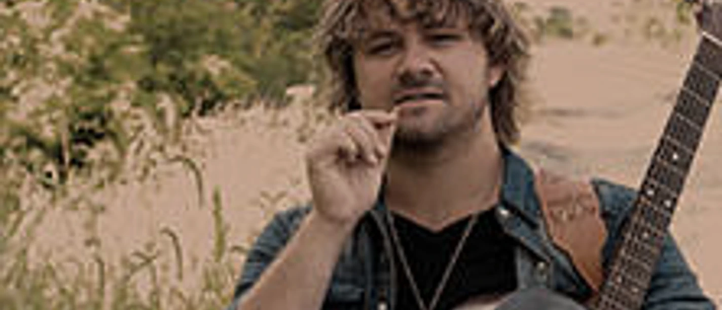 Mann mit Gitarre steht draußen vor Vegetation, hält ein Grashalmstück zwischen den Fingern.