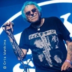 Älterer Musiker mit Mikrofon und Sonnenbrille auf einer Bühne vor blauem Hintergrund