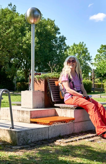 Mülheimer RUHRPERLEN Frau sitzt bei sonnigem Wetter fröhlich auf einer Bank an einer Säule (RUHRPERLE), umgeben von Natur und wenigen parkenden Autos