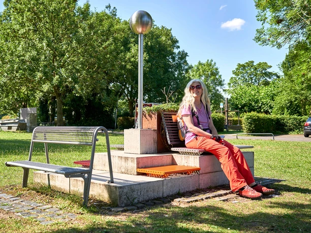Mülheimer RUHRPERLEN Frau sitzt bei sonnigem Wetter fröhlich auf einer Bank an einer Säule (RUHRPERLE), umgeben von Natur und wenigen parkenden Autos
