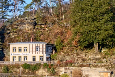 Haus mit Fachwerk und Schild "Heimatmuseum & Pflanzgarten" vor bewaldetem Felsabbruch.