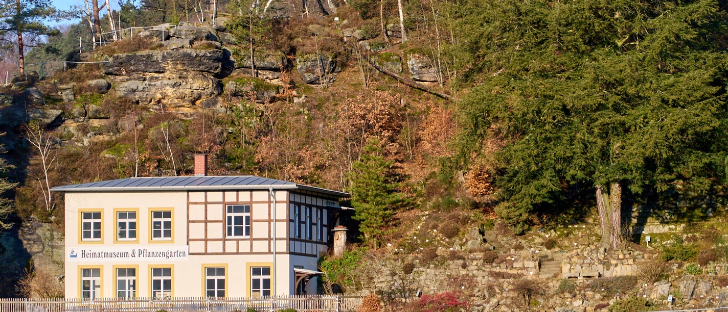 Haus mit Fachwerk und Schild "Heimatmuseum & Pflanzgarten" vor bewaldetem Felsabbruch.