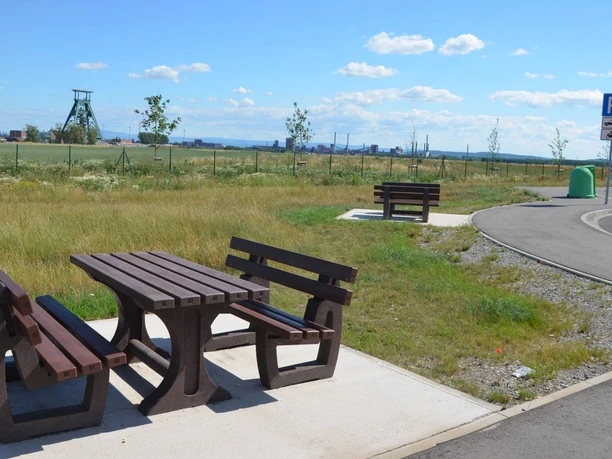 Holztisch mit Bänken an einer Raststätte neben Parkplatz, im Hintergrund Wiesen und Industriebauten.