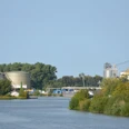 Salzgitter-Beddingen_Hafen (24)_HD_16zu9.JPG Industriehafen mit Kränen, Silos und Lagergebäuden am Wasser, umgeben von Bäumen und Grünflächen.