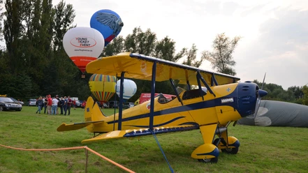Flugtage_Salzgittersee (5)_HD_16zu9.JPG Gelbes Doppeldeckerflugzeug auf einer Wiese, dahinter bunte Heißluftballons und Besuchergruppen.
