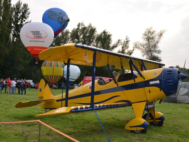 Flugtage_Salzgittersee (5)_HD_16zu9.JPG Gelbes Doppeldeckerflugzeug auf einer Wiese, dahinter bunte Heißluftballons und Besuchergruppen.