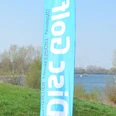 Blaue Disc-Golf-Fahne am Ufer des Salzgittersees vor Bäumen, Wasser und Windrädern im Hintergrund.