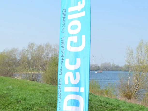 Disc-Golf_Salzgittersee (24)_HD_16zu9.JPG Blaue Disc-Golf-Fahne am Ufer des Salzgittersees vor Bäumen, Wasser und Windrädern im Hintergrund.