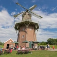Mühlencafé Holtlander Mühle Menschen sitzen vor einer großen Windmühle. Du siehst Tische, Sonnenschirme und blauen Himmel.