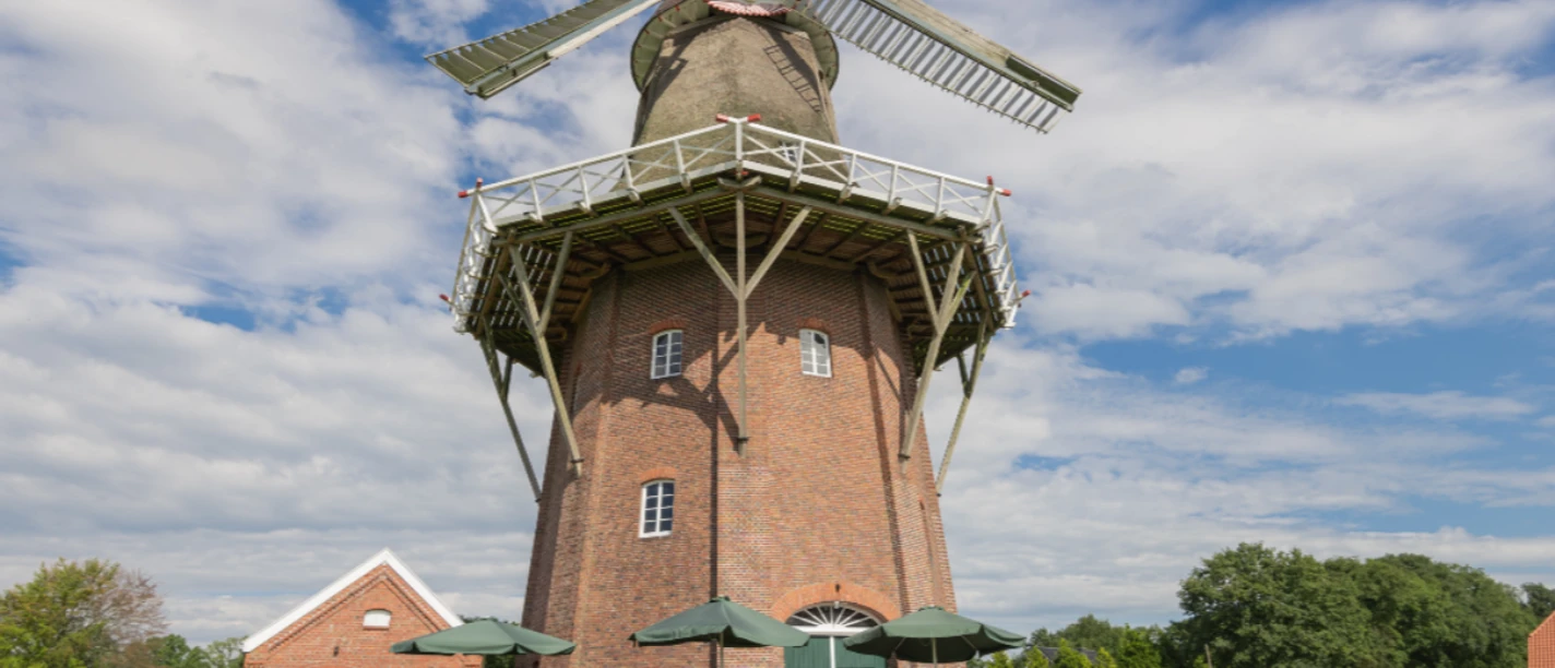 Mühlencafé Holtlander Mühle Menschen sitzen vor einer großen Windmühle. Du siehst Tische, Sonnenschirme und blauen Himmel.