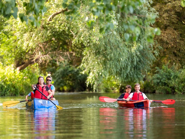 WF-Wassersport-05.jpg Menschen paddeln in farbigen Kanus auf einem ruhigen Fluss, umgeben von dichtem grünen Laub.