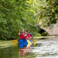 Zwei Erwachsene und ein Kind paddeln im Kanu auf einem Fluss vor einer Steinbrücke