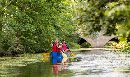 WF-Wassersport-08.jpg Zwei Erwachsene und ein Kind paddeln im Kanu auf einem Fluss vor einer Steinbrücke