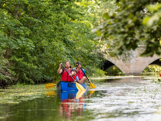 WF-Wassersport-08.jpg Zwei Erwachsene und ein Kind paddeln im Kanu auf einem Fluss vor einer Steinbrücke