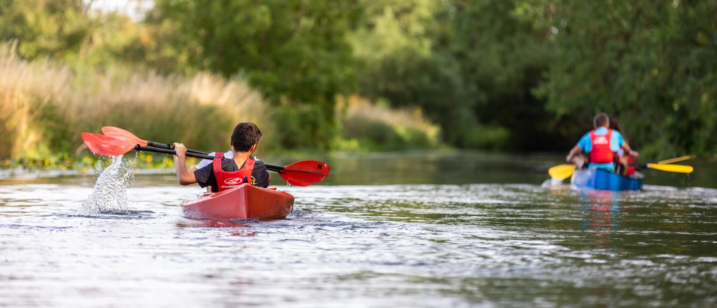 WF-Wassersport-04.jpg Zwei Kanus mit Paddlern auf einem ruhigen Fluss, umgeben von grüner Ufervegetation.