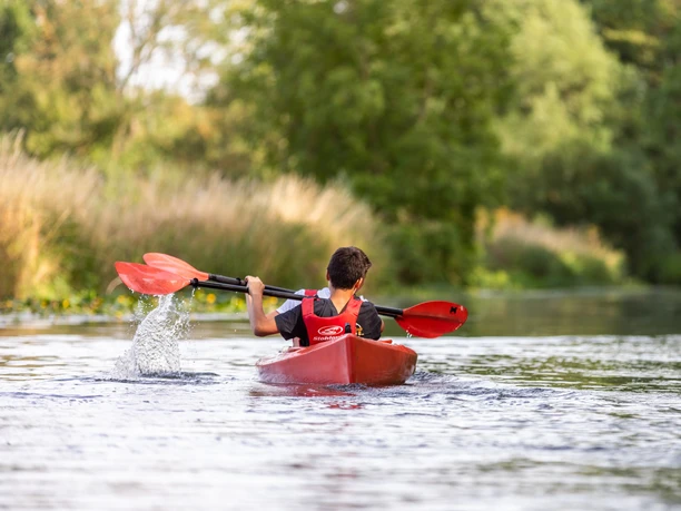 WF-Wassersport-04.jpg Zwei Kanus mit Paddlern auf einem ruhigen Fluss, umgeben von grüner Ufervegetation.