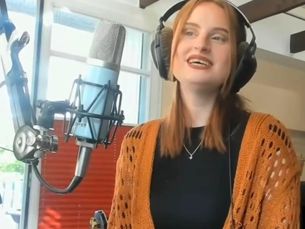 Künstlerin.jpg Frau mit langen Haaren trägt Kopfhörer und steht vor einem großen Mikrofon in einem Raum mit Holzbalken an der Decke