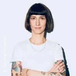 Frau mit kurzem Bob, weißem T-Shirt und tätowierten Armen steht mit verschränkten Armen.