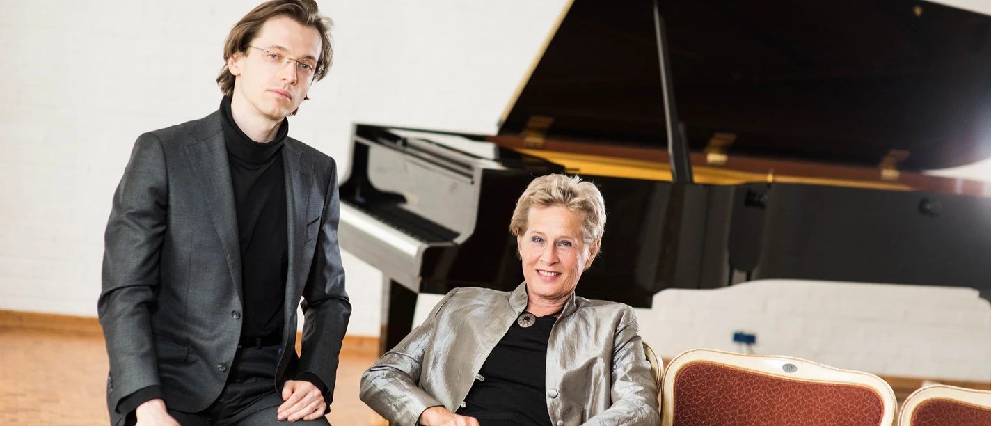 erik_satie_klein.jpg Zwei Musiker in eleganter Kleidung sitzen vor einem Konzertflügel in einem hellen Saal.