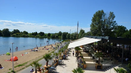 Salzgitter_Cafedellago_HD_16zu9.JPG Restaurantterrasse am Seeufer mit Gästen, Strandbereich und Badenden unter blauem Himmel