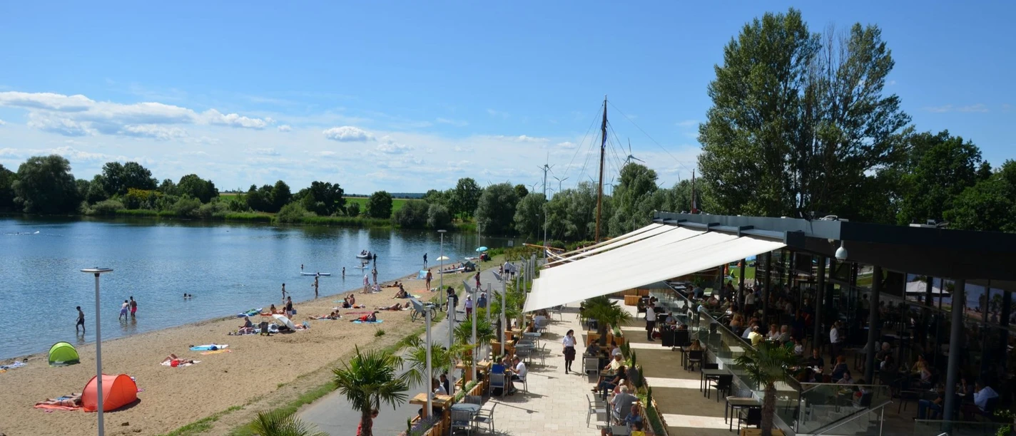 Salzgitter_Cafedellago_HD_16zu9.JPG Restaurantterrasse am Seeufer mit Gästen, Strandbereich und Badenden unter blauem Himmel