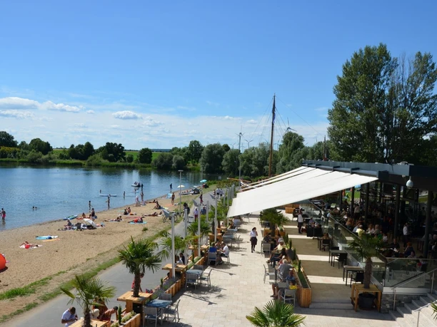 Salzgitter_Cafedellago_HD_16zu9.JPG Restaurantterrasse am Seeufer mit Gästen, Strandbereich und Badenden unter blauem Himmel
