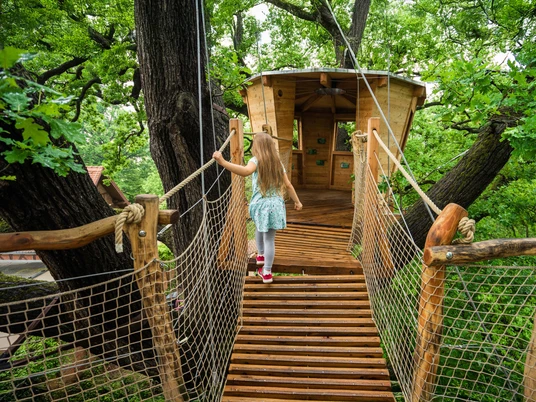 Beobachtungsplattform der Auwaldstation Leipzig - Sehenswürdigkeiten in Leipzig Ein Mädchen läuft über eine hölzerne Hängebrücke zur Beobachtungsplattform der Auwaldstation Leipzig in den Baumkronen, die wie ein Baumhaus aussieht.