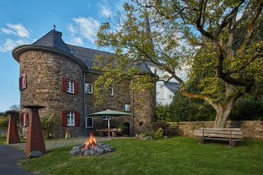 Burg Honrath mit Rundturm, roten Fensterläden, Feuerstelle und Gartenbank unter einem Baum