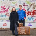 Theaterplakat zu Leonce und Lena Zwei Personen stehen vor einer mit Graffiti bemalten Wand. Zwei alte Lederkoffer stehen vor ihren Füßen.Two people are standing in front of a wall painted with graffiti. Two old leather suitcases stand at their feet.
