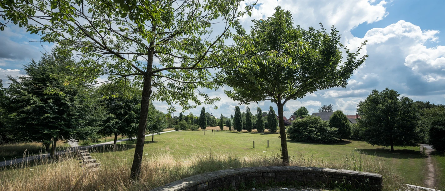 Park Kattenstroth Blick über Wiese mit Bäumen, gepflasterte Terrasse im Vordergrund, Weg und Häuser am Rand.