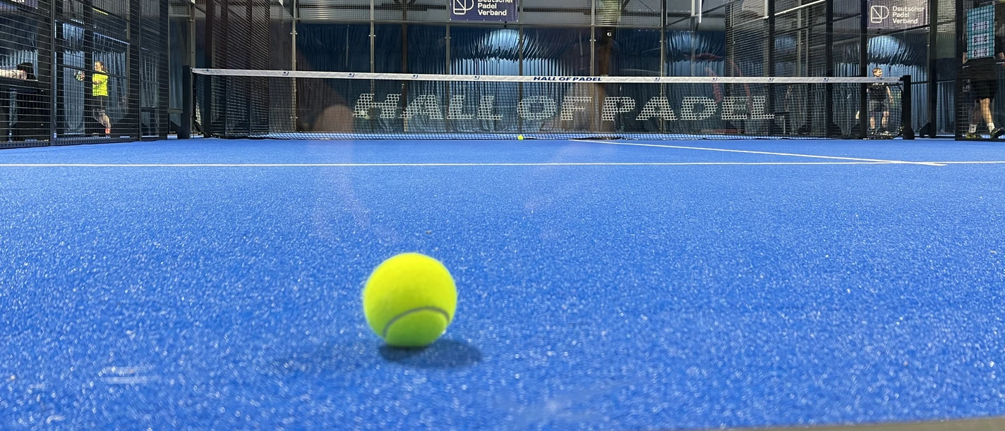 Hall of Padel Padelplatz mit blauem Boden und Netz in überdachter Halle, im Vordergrund ein gelber Ball.