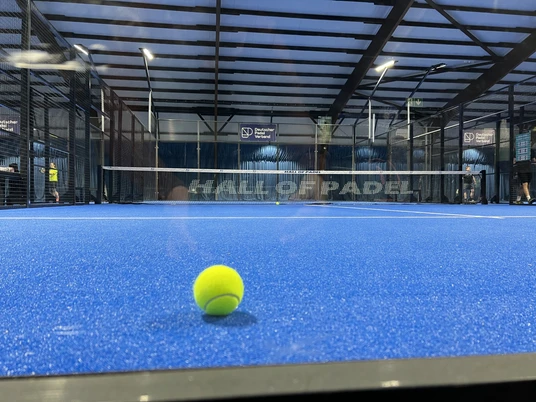 Hall of Padel Padelplatz mit blauem Boden und Netz in überdachter Halle, im Vordergrund ein gelber Ball.