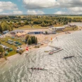 ALL-on-SEA Camp & Sport Resort am Schladitzer See - Leipziger Neuseenland Blick auf die Schladitzer Bucht im Leipziger Neuseenland mit dem All-on-Sea Camp & Sport Resort. Im Sand und Gras liegen Kanus und Kajaks, dahinter befinden sich die Gastronomie mit überdachten Sitzplätzen im Freien sowie das Beachvolleyballfeld. Im Wasser sind zwei Rudelboote zu sehen.
