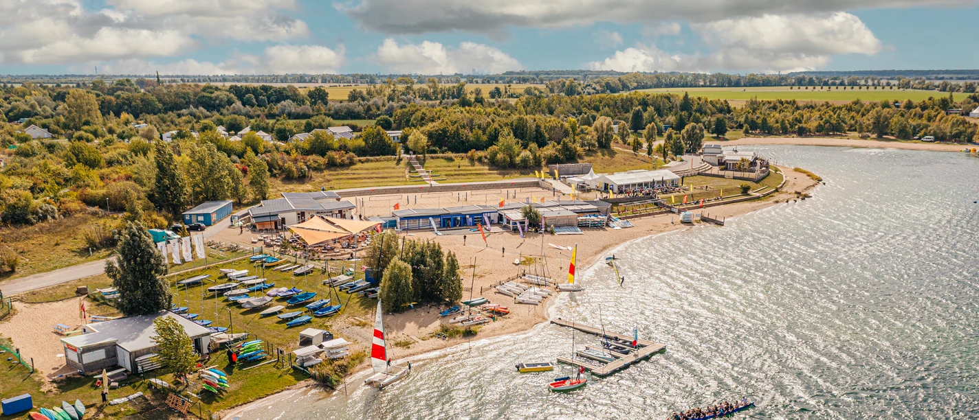 ALL-on-SEA Camp & Sport Resort am Schladitzer See - Leipziger Neuseenland Blick auf die Schladitzer Bucht im Leipziger Neuseenland mit dem All-on-Sea Camp & Sport Resort. Im Sand und Gras liegen Kanus und Kajaks, dahinter befinden sich die Gastronomie mit überdachten Sitzplätzen im Freien sowie das Beachvolleyballfeld. Im Wasser sind zwei Rudelboote zu sehen.