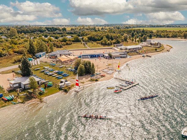 Vue sur la baie de Schladitz dans le Leipziger Neuseenland avec le All-on-Sea Camp & Sport Resort. Des canoës et des kayaks sont posés dans le sable et l'herbe, derrière lesquels se trouvent la gastronomie avec des places assises couvertes en plein air ainsi que le terrain de beach-volley. Dans l'eau, on peut voir deux bateaux de meute.