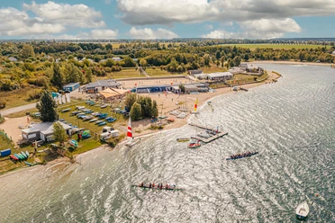 Widok na zatokę Schladitz w lipskim Neuseenland z All-on-Sea Camp & Sport Resort. Kajaki i kanoe leżą na piasku i trawie, za nimi znajduje się zaplecze gastronomiczne z zadaszonymi miejscami do siedzenia na zewnątrz i boisko do siatkówki plażowej. W wodzie można zobaczyć dwie łodzie.