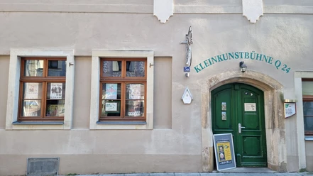 Hauswand mit zwei Fenstern und einer grünen Tür. Über dem Türeingang steht Kleinkunstbühne Q24