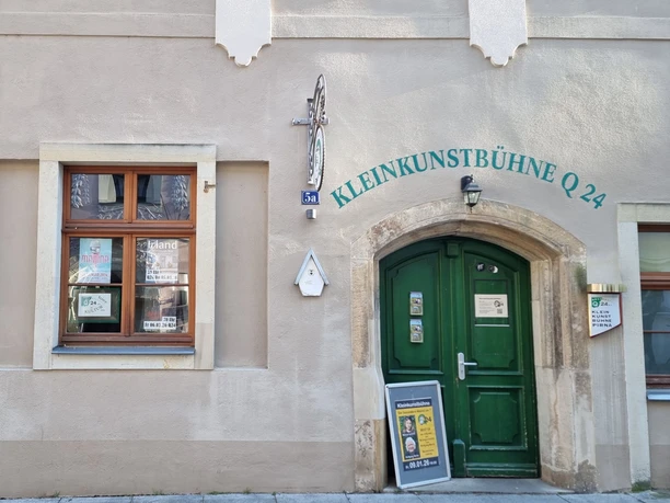 Hauswand mit zwei Fenstern und einer grünen Tür. Über dem Türeingang steht Kleinkunstbühne Q24