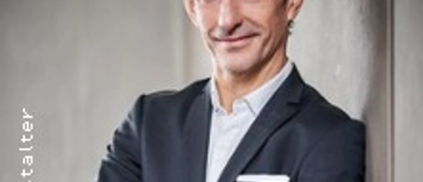 Ein Mann im Anzug lehnt an einer Wand und blickt mit verschränkten Armen in die Kamera.