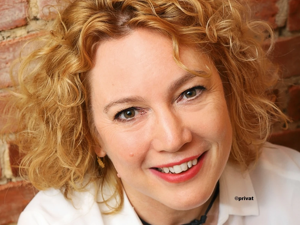 Salhofen_Maike-1.jpg Person mit lockigem, schulterlangem Haar vor einer Ziegelsteinwand trägt ein weißes Hemd und eine Halskette mit Anhänger