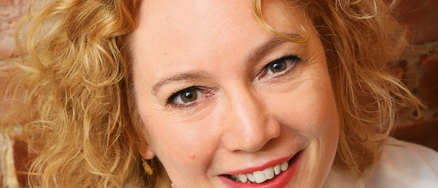 Salhofen_Maike-1.jpg Person mit lockigem, schulterlangem Haar vor einer Ziegelsteinwand trägt ein weißes Hemd und eine Halskette mit Anhänger