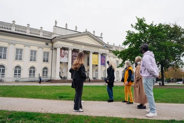 Stadtrundgang Kassel-Kunst-Kultur Auf dem Friedrichsplatz steht eine Gruppe vor dem Museum Fridericianum und hört einem Gästeführer zu.