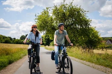 Radfahren im Osnabrücker Land Zwei Radfahrende unterwegs auf einem ruhigen Weg im Osnabrücker Land, umgeben von Feldern, Bäumen und offener Landschaft.