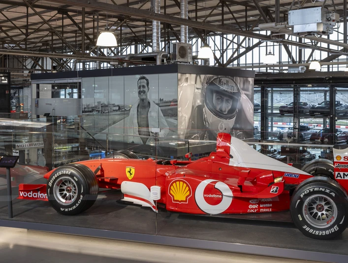 Michael Schumacher Private Collection Roter Formelwagen in verglaster Ausstellungshalle mit großformatigen Fotos von Michael Schuhmacher im HintergrundRed formula car in glazed exhibition hall with large-format photos of Michael Schuhmacher in the background