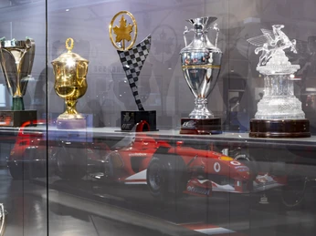 Michael Schumacher Private Collection Vitrine mit Rennsportpokalen vor dem Spiegelbild eines roten Formelwagens.Display case with racing trophies in front of the reflection of a red formula car.