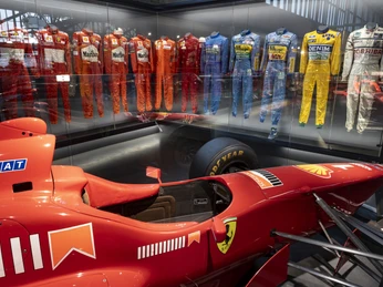 Michael Schumacher Private Collection Rotes Formelauto vor einer Glaswand mit farbigen Rennanzügen in einer Ausstellung.Red formula car in front of a glass wall with colored racing suits in an exhibition.