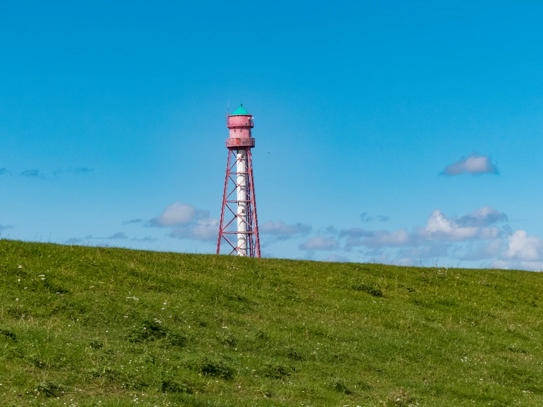 Deich beim Campener Leuchtturm Roter Stahlturm mit grünem Dach ragt hinter einer grasbewachsenen Deichlandschaft in den blauen Himmel.