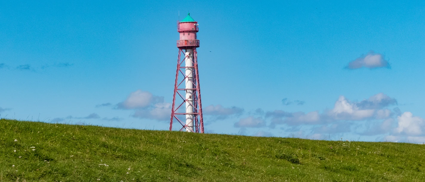 Deich beim Campener Leuchtturm Roter Stahlturm mit grünem Dach ragt hinter einer grasbewachsenen Deichlandschaft in den blauen Himmel.